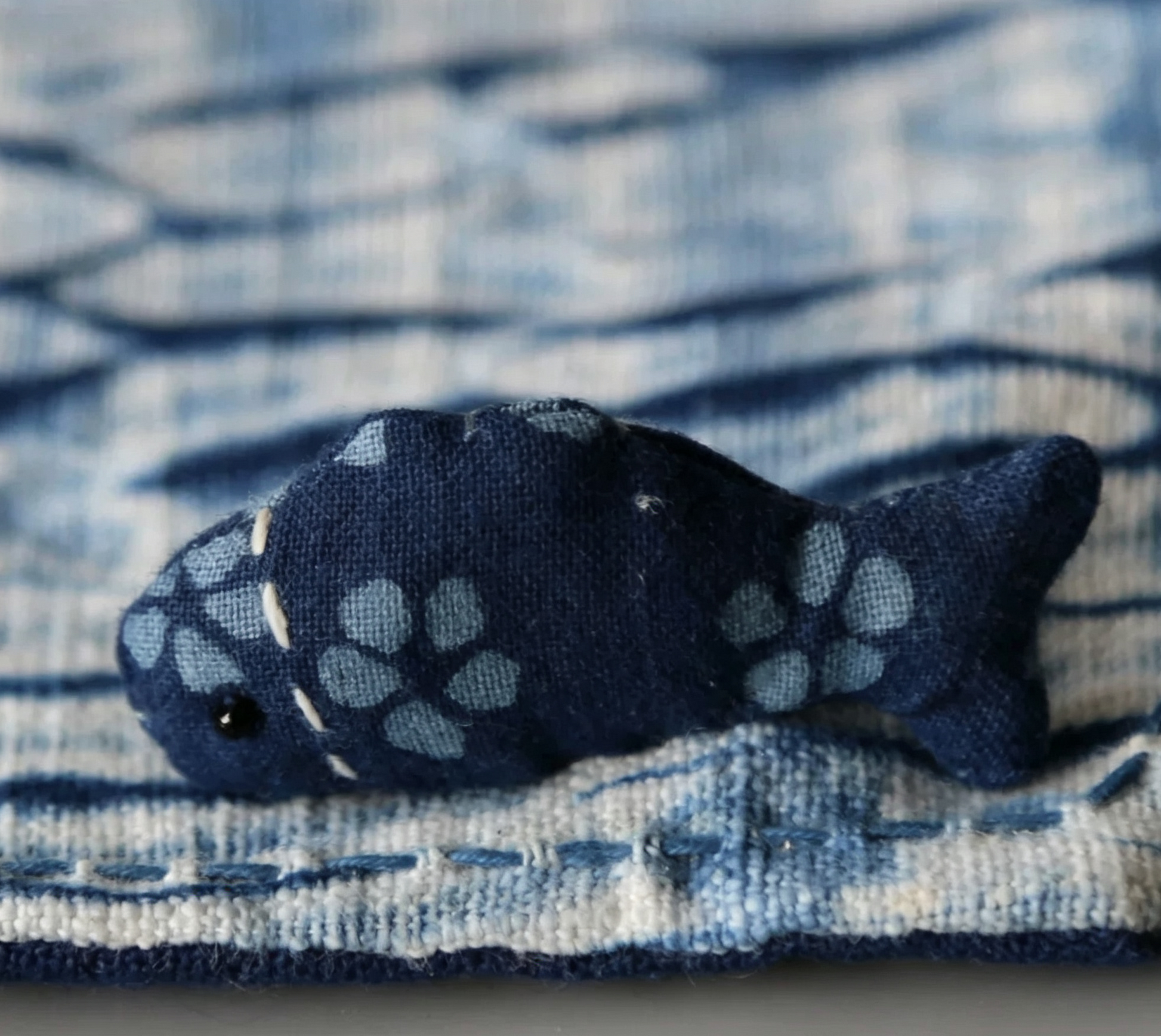 Macro detail of a hand-sewn fish charm on a botanical indigo dyed textile, showcasing artisanal craftsmanship and texture.