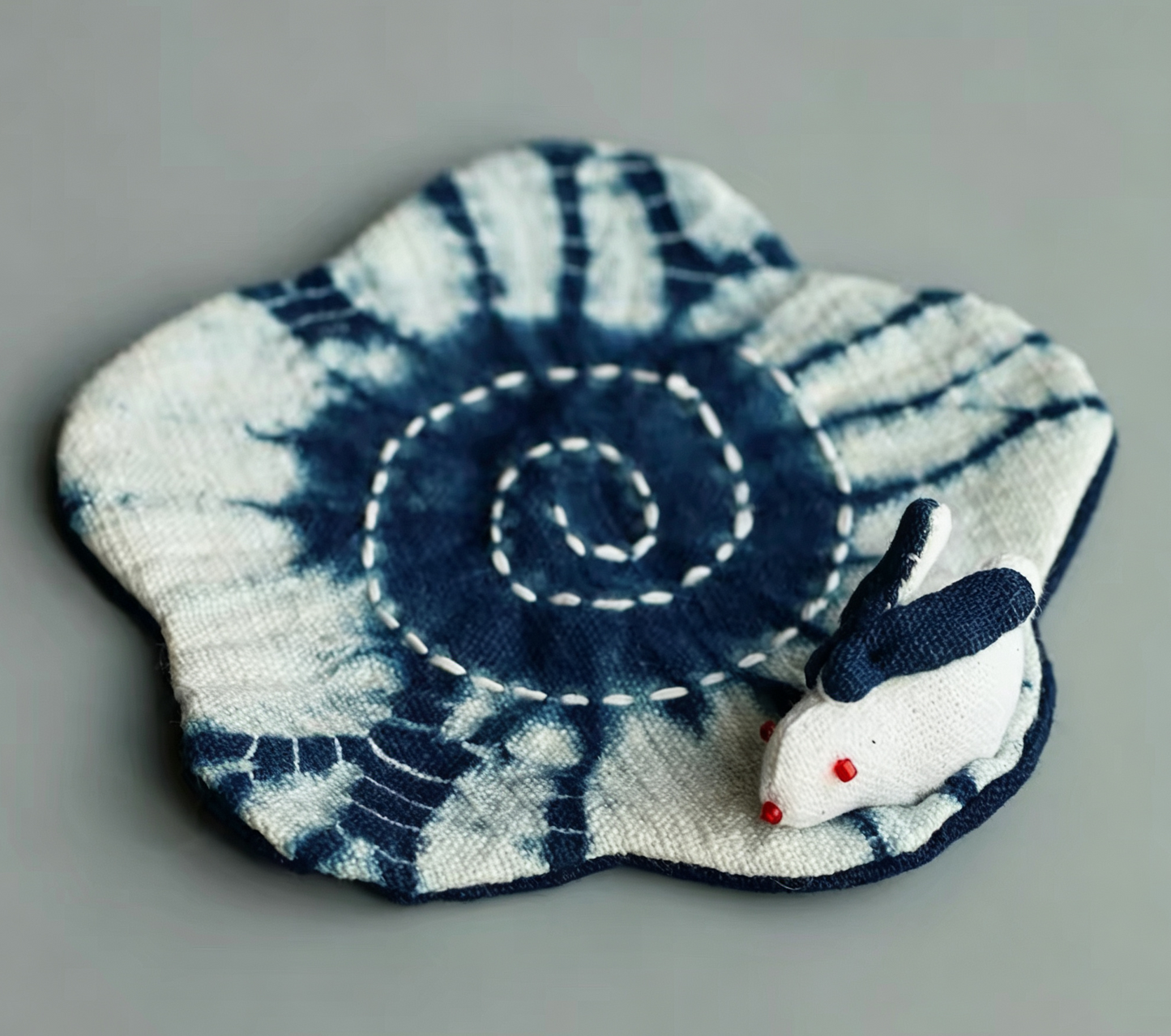 Top view of an artisanal rabbit-shaped indigo mat featuring a white hand-stitched bunny on a spiraling tie-dye cotton base.