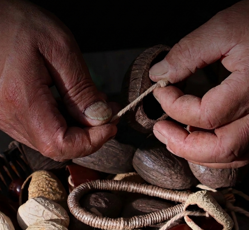 Close-up of artisan hands meticulously threading natural fruit shells with hemp rope, highlighting the handcrafted process of the percussion instrument