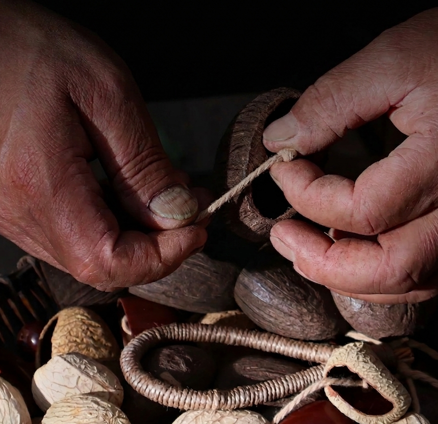 Close-up of artisan hands meticulously threading natural fruit shells with hemp rope, highlighting the handcrafted process of the percussion instrument