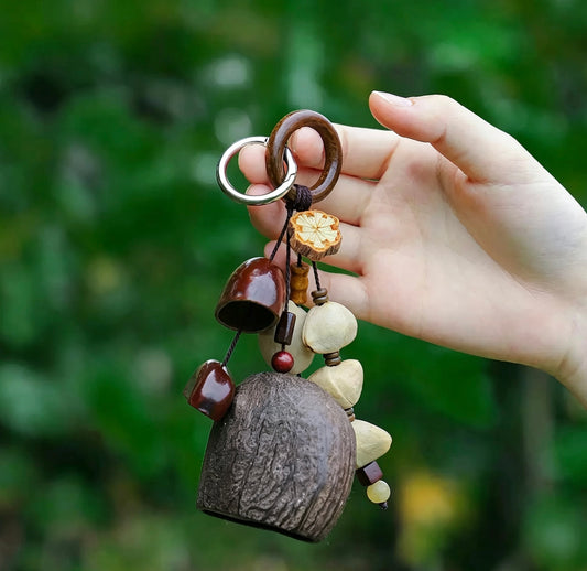 Close-up of a hand holding a handcrafted nut shell shaker charm. It showcases a large dark brown Salin shell, polished red wooden beads, Pala,Chacha seeds, and a floral-patterned disc, set against a bright green natural background.