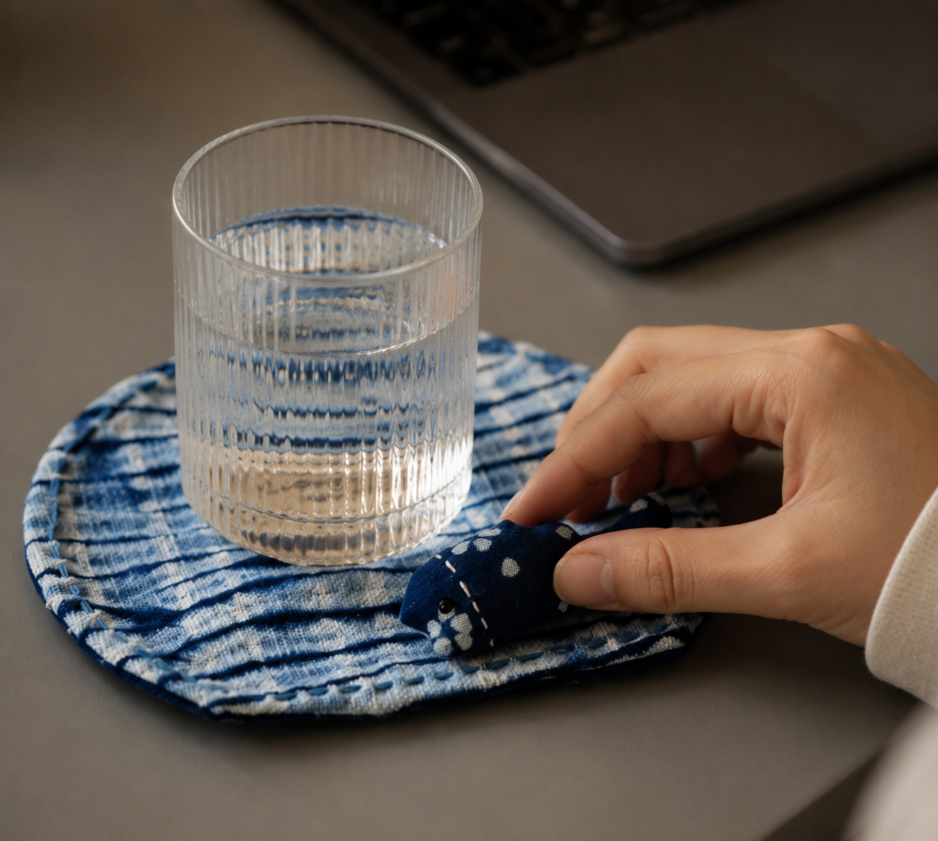 A glass of water on a hand-sewn indigo desktop mat, a hand touching the fabric companion next to a laptop, mindful workspace setting.