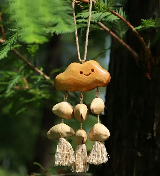 Close-up of a handmade "Rainy Cloud" wooden hanging ornament suspended from a tree branch, featuring a smiling face design, natural shell droplets, and hemp tassels.