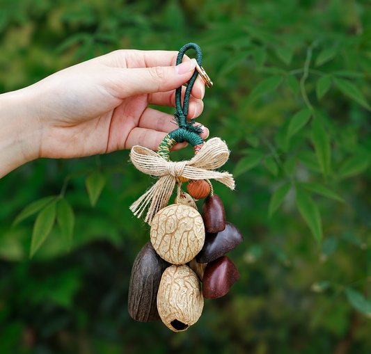 Close-up of a hand holding a handmade nut shell shaker charm. The image highlights the “five-color" woven loop, jute bow, and a combination of kinds of dried shells, set against a blurred leafy background