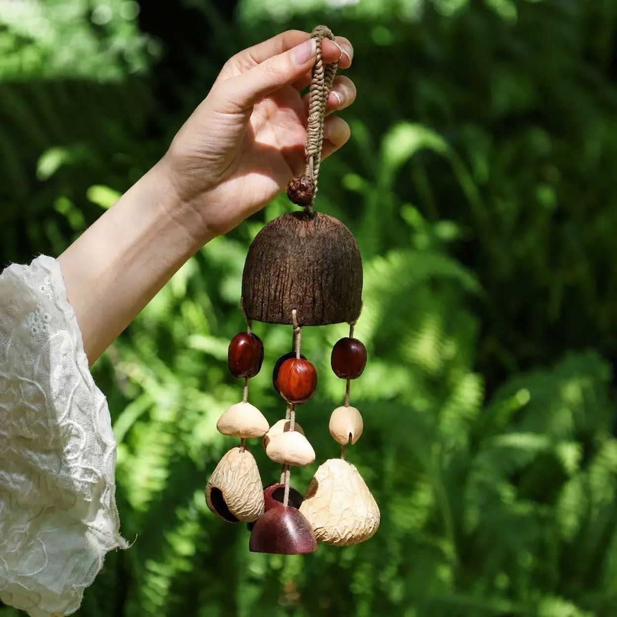 A hand holding a large handmade natural wind chime with a dark Salin shell top, featuring polished Pala shells and raw white Chacha and Panji  hanging below, set against a lush green garden background.